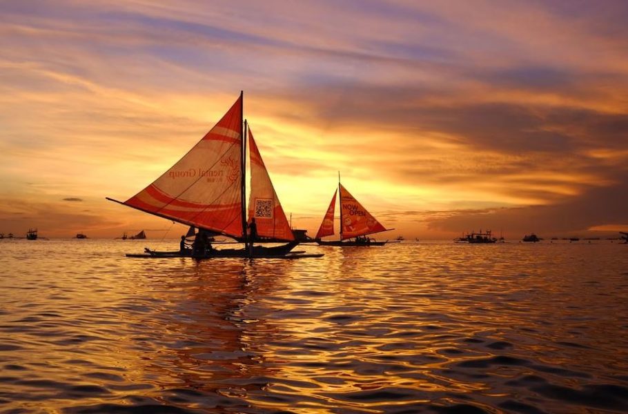 Dramatic sunset over White Beach Boracay with silhouetted paraw sailing boats lined up along shore, orange and purple clouds reflected in wet sand, person running on beach